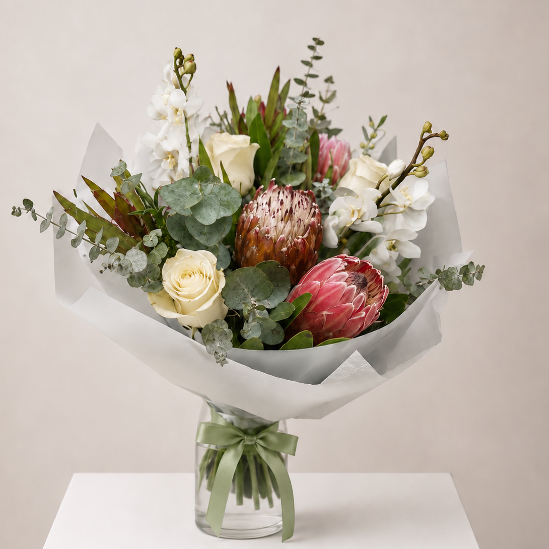 A floral bouquet in a clear glass vase with a light green ribbon, displayed on a white pedestal against a warm beige background. The bouquet features white roses, white orchids, pink protea, green eucalyptus, and a textured Native Florals. 